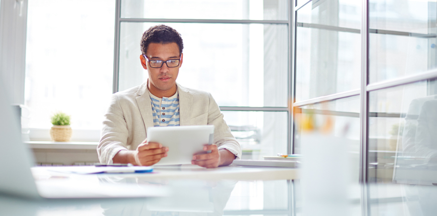 African American man sits at a table in an office environment, reading off a tablet.
