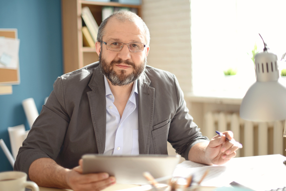 An older, bearded man sits at a desk, tablet in hand.